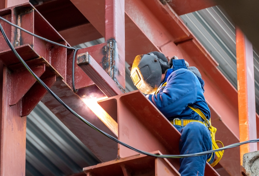 事業内容「鍛冶工事」のイメージ。高所で鉄骨に溶接作業を行う作業員の様子。溶接面を着用し、安全帯を装着した姿が確認できる。