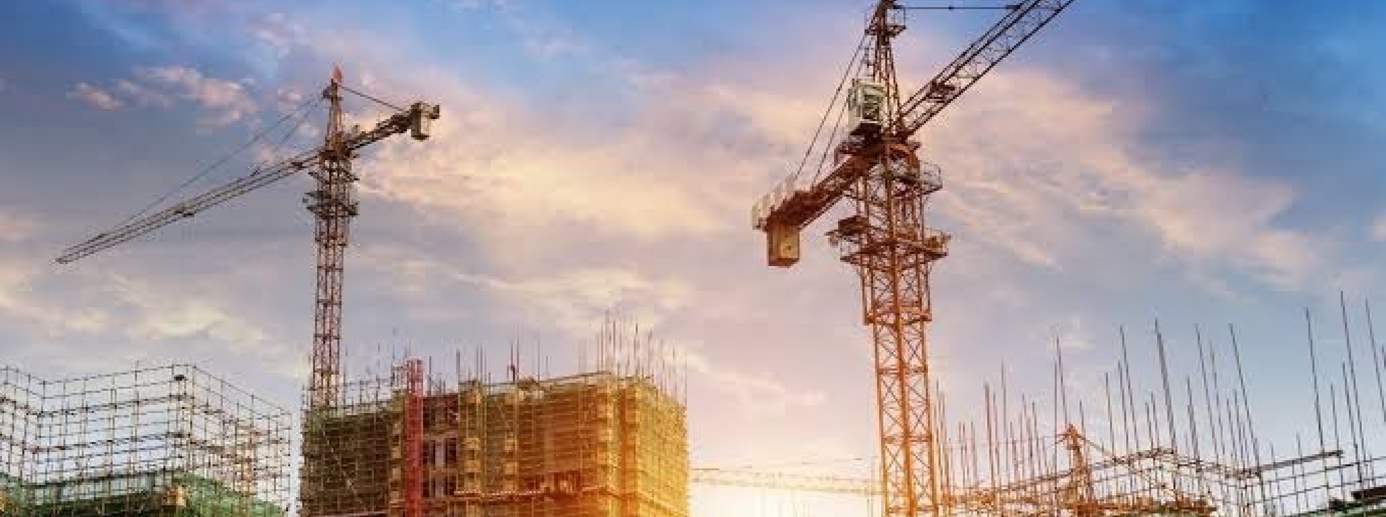 夕空の下、複数のタワークレーンが稼働し、足場が組まれた建設中のビル群が並ぶ工事現場の風景。